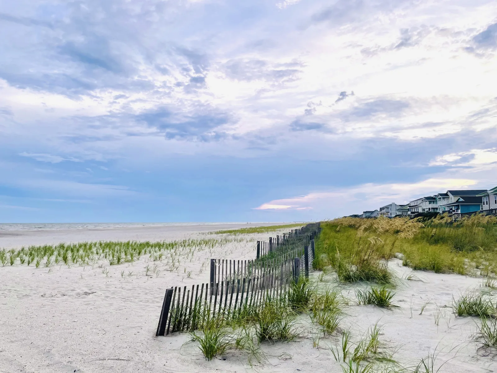 A quiet North Carolina beach at dusk, with a weathered sand fence, dune grass and beachfront houses along the shore